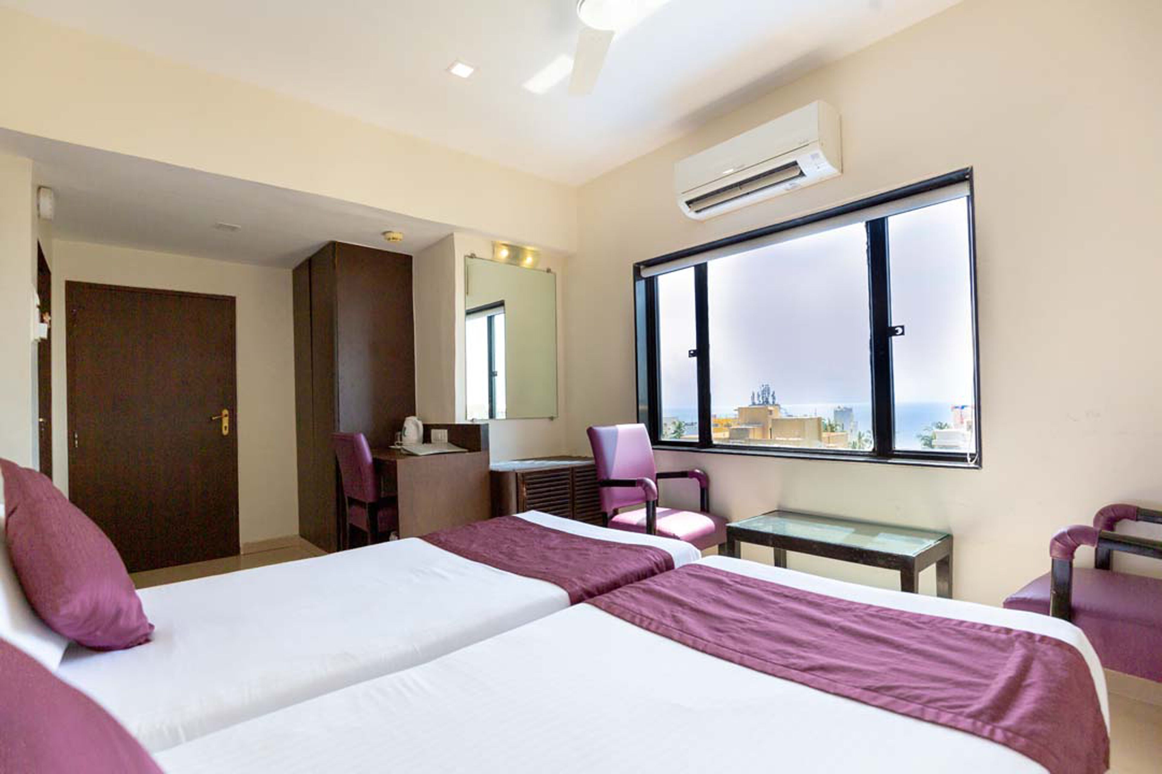 A hotel room with two beds, a desk, a chair, and a large window with a city view.