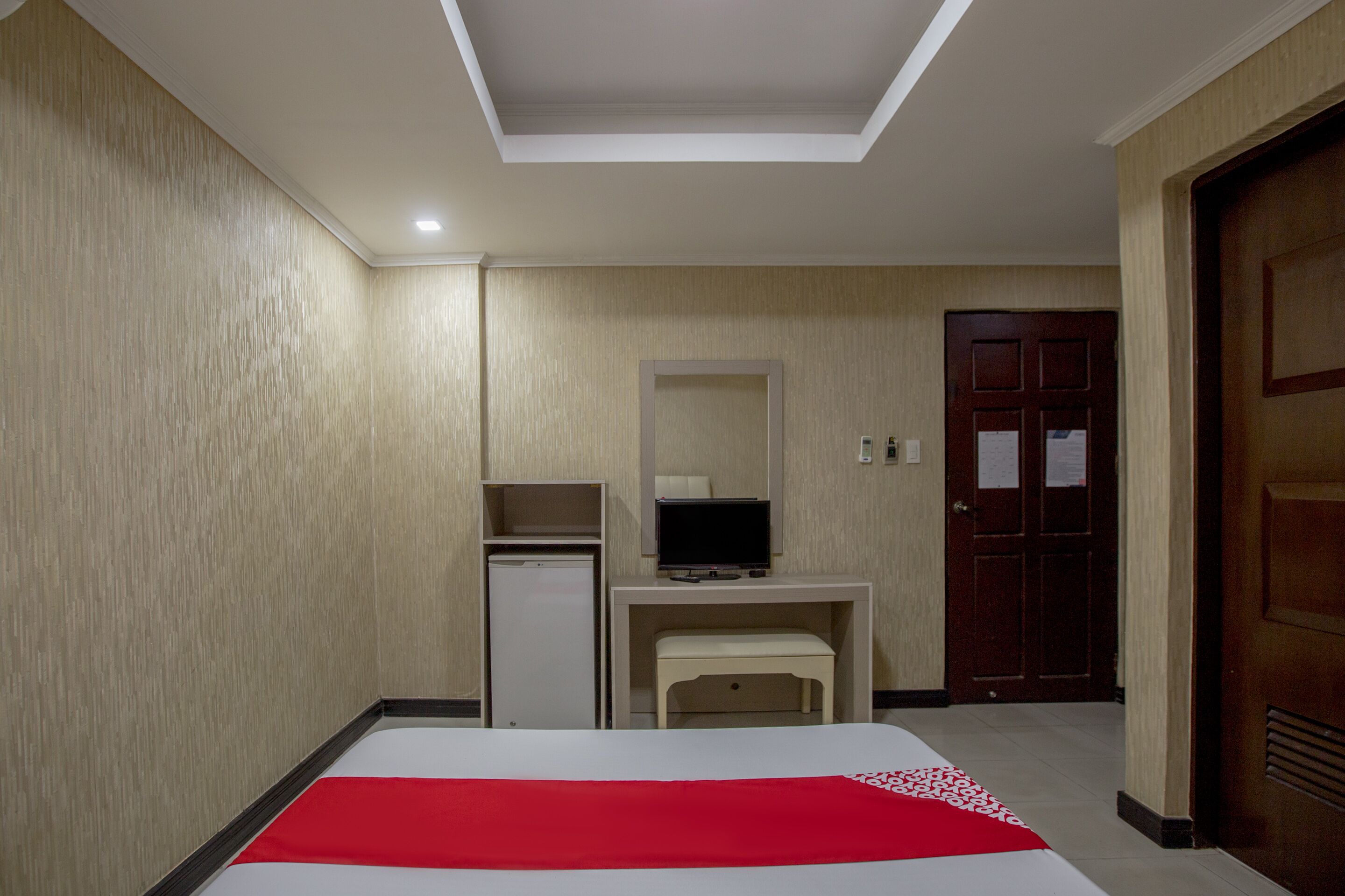 A hotel room with a bed, a desk with a television, a small refrigerator, and a door.
