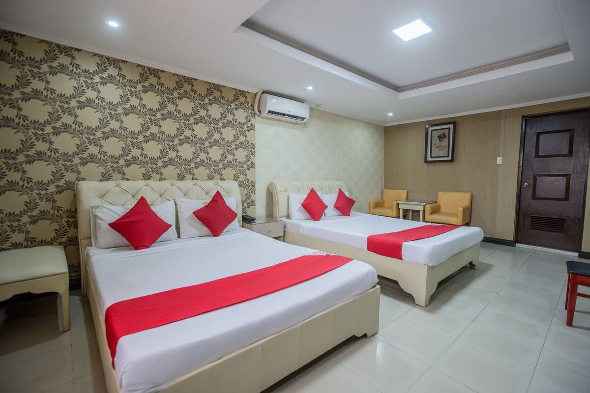A hotel room with two beds, a small table, a chair, and an air conditioning unit on the wall.