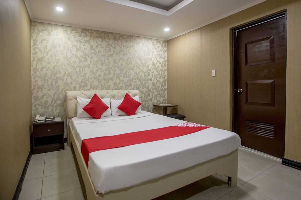A neatly made bed with white and red bedding, a bedside table with a phone and a book, and a wooden door in a room with patterned wallpaper.