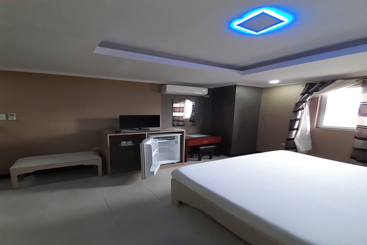 A hotel room with a bed, a desk, a chair, a bench, an air conditioning unit, and a ceiling light.