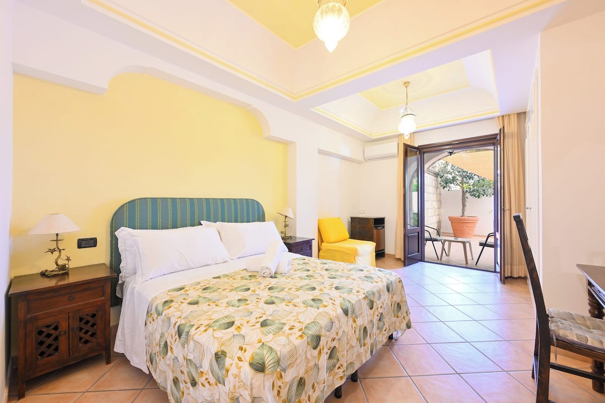 A hotel room with a bed, a nightstand, a chair, a table, and a balcony with a potted plant.