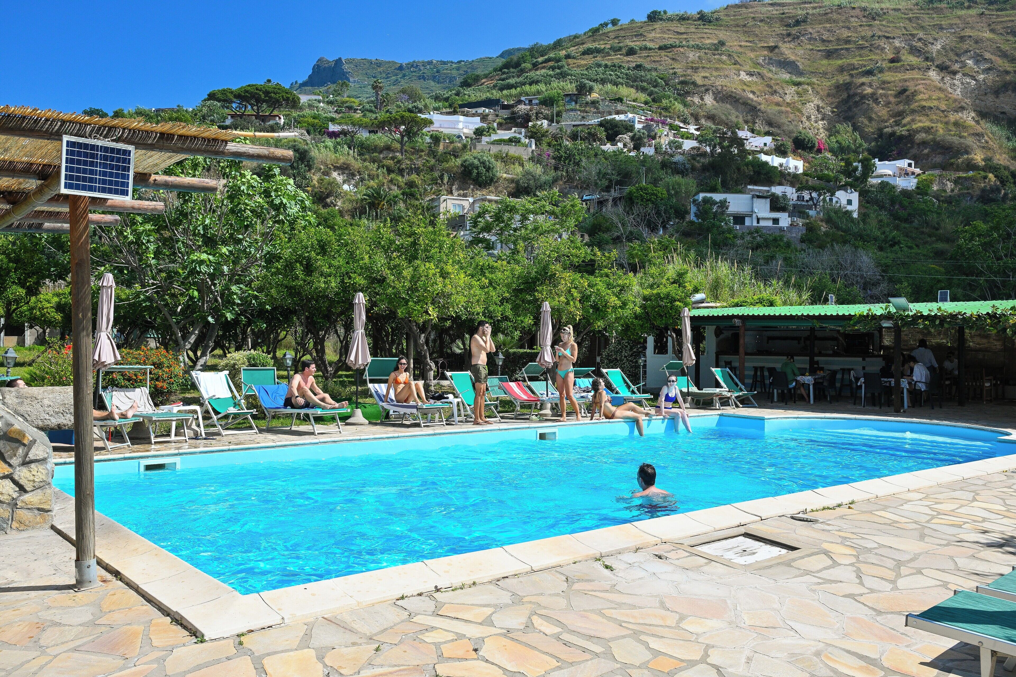 Une piscine entourée de chaises longues et de parasols, bordée de dalles de pierre et de verdure, avec une colline en arrière-plan.