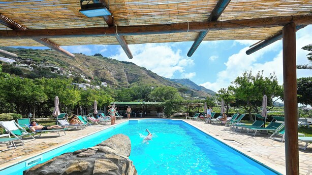 A swimming pool with lounge chairs and umbrellas, surrounded by greenery and mountains. - Villa Tara (Serrara Fontana)
