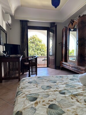 A hotel room with a bed, a desk with a television, a chair, and a balcony with a view of greenery. - Villa Tara (Serrara Fontana)