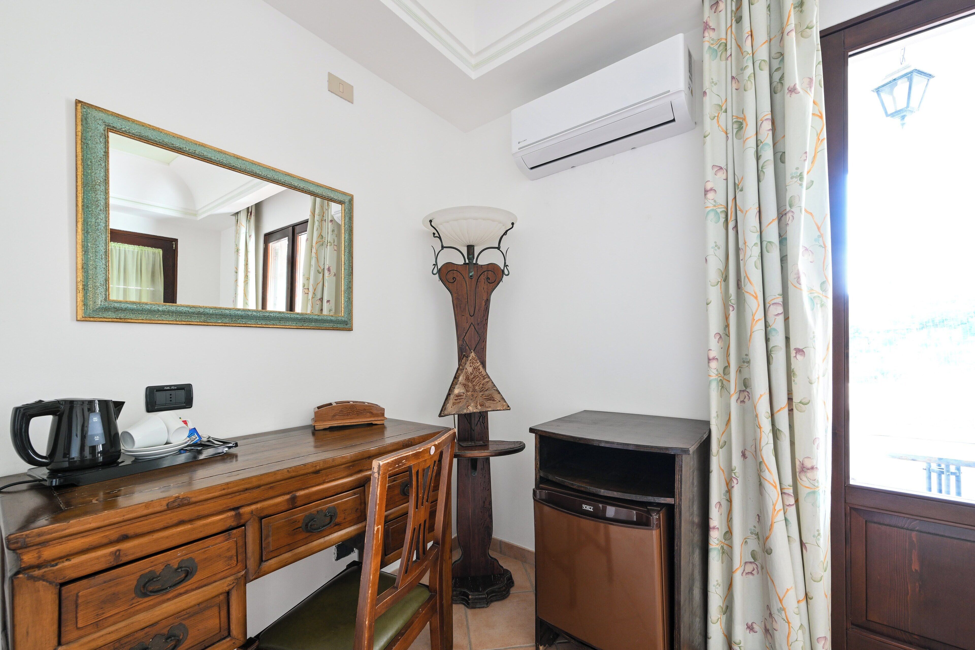 Una habitación con un escritorio de madera, un espejo, una lámpara de pie y un ventanal con cortinas.