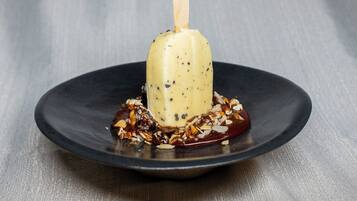 Ein Dessert auf einem schwarzen Teller, mit einem Holzspieß, umgeben von Nüssen und Schokoladensauce.