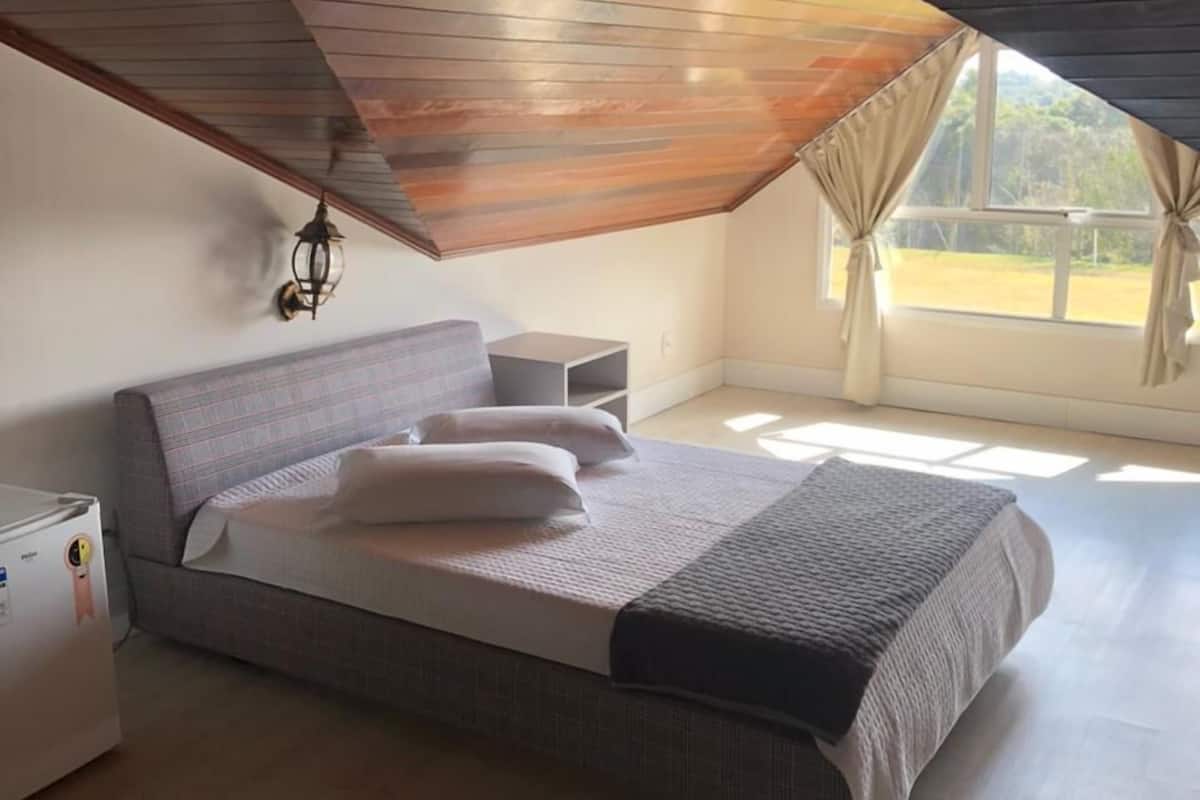 Un dormitorio con techo inclinado, una cama, una mesita de noche, una lámpara y un ventanal con cortinas.
