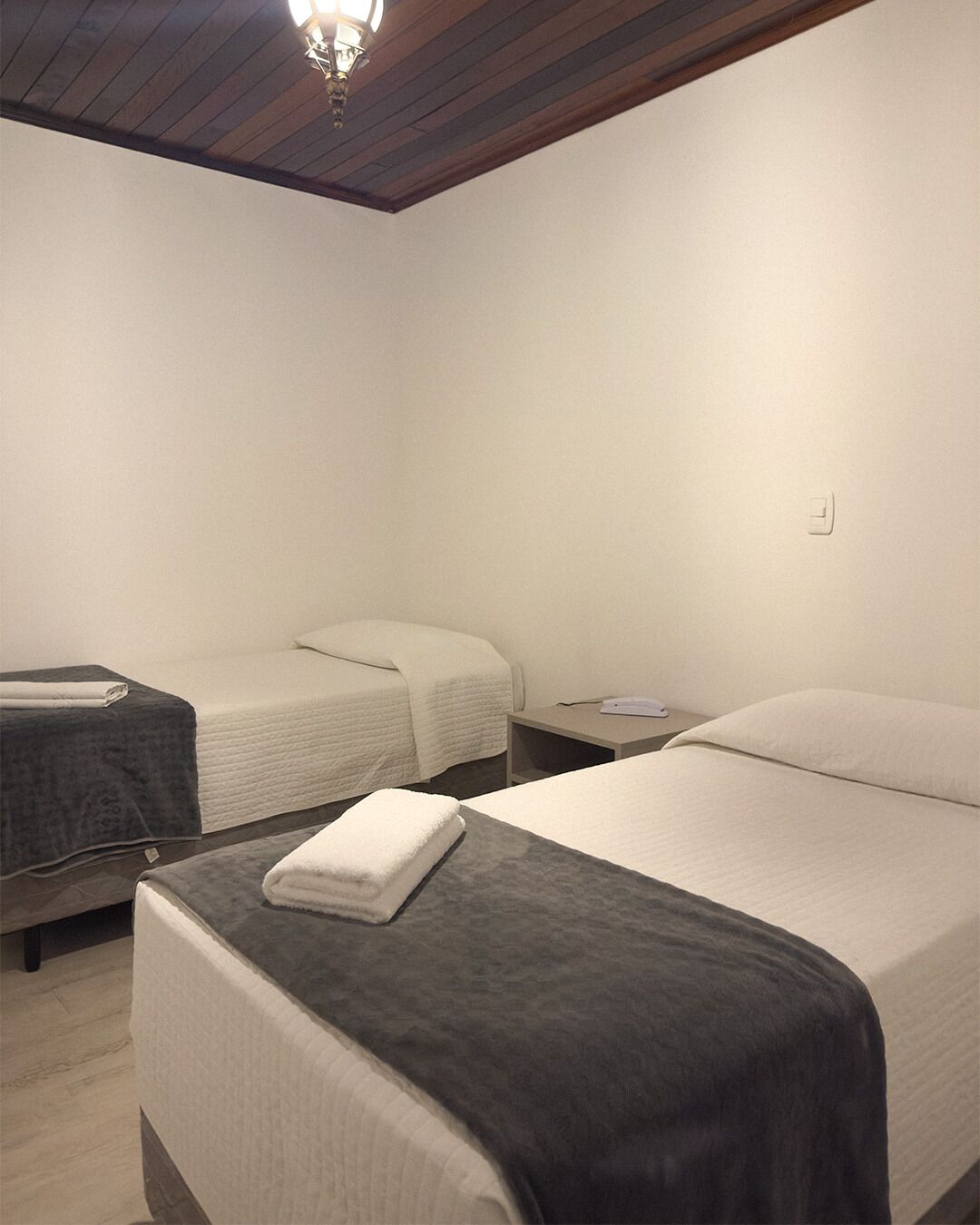 A hotel room with two beds, each with a white bedspread and a dark blanket. There is a small bedside table with a lamp and a power outlet on the wall.