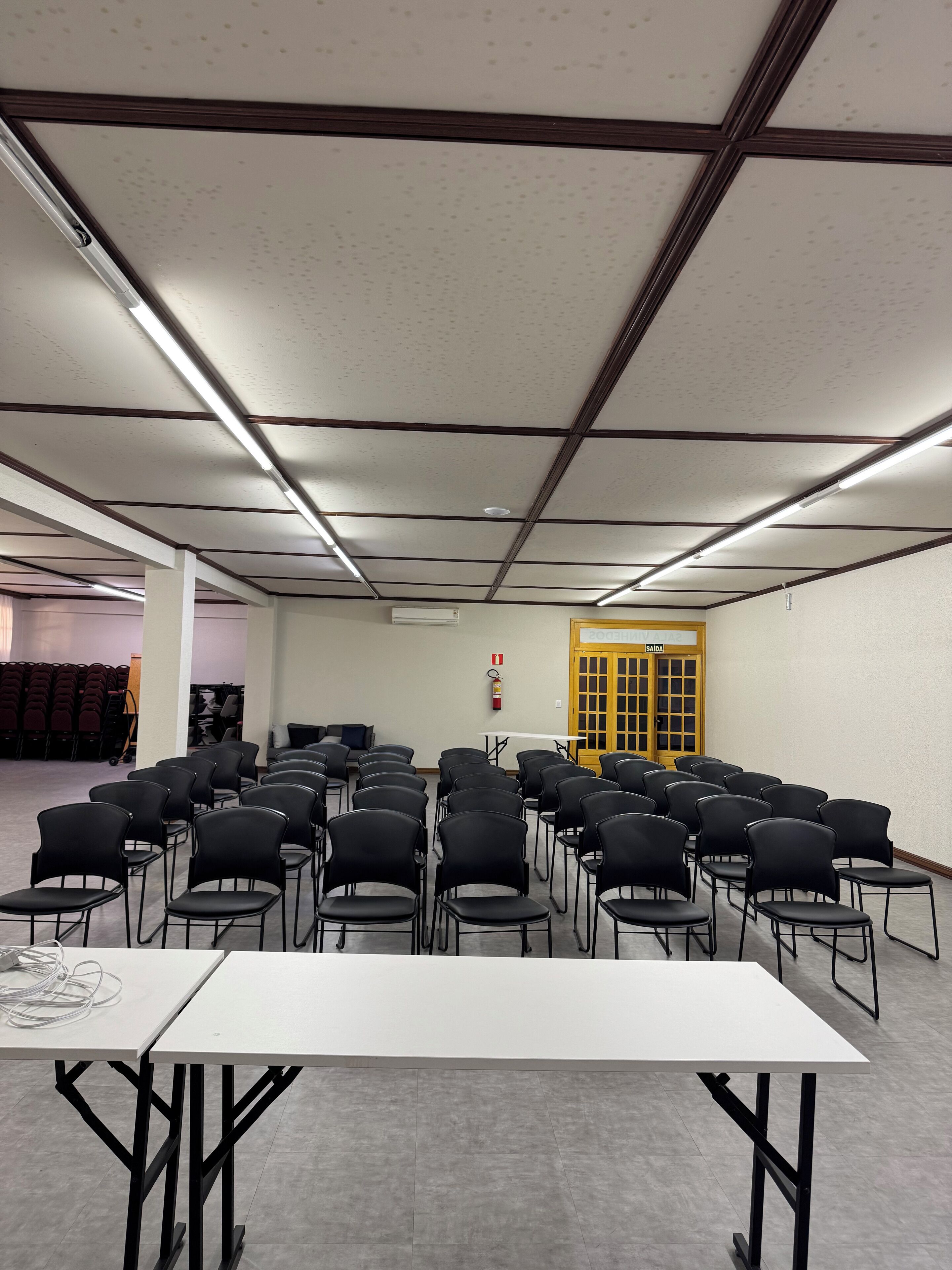 Une salle de conférence avec des rangées de chaises noires, une table blanche et un plafond doté d’un éclairage fluorescent.