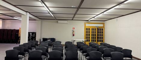 Une salle de conférence avec des rangées de chaises noires, une table blanche et un plafond doté d’un éclairage fluorescent.