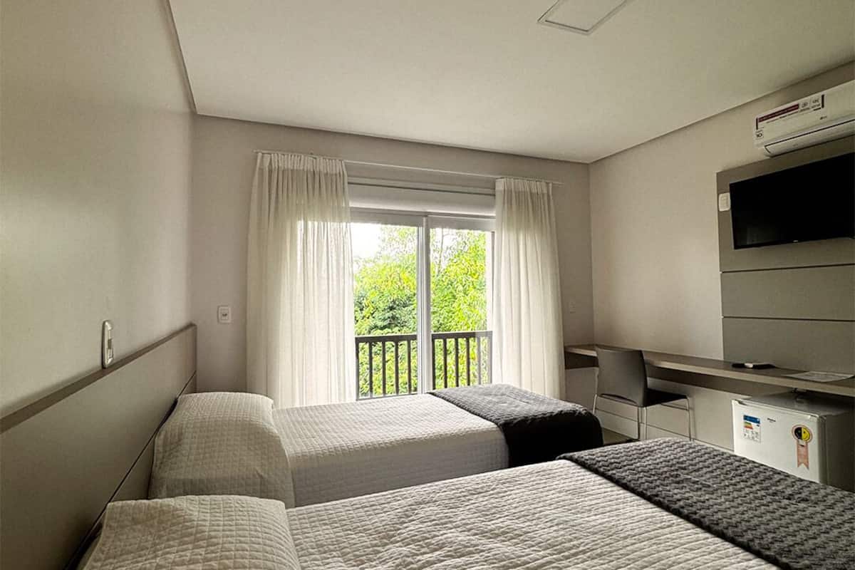 Habitación de hotel con dos camas, televisión, escritorio y balcón con cortinas.