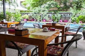 A poolside area with wicker furniture and a dining table, surrounded by lush greenery and a pool. - The 252 (Phnom Penh)