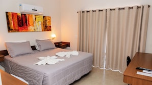 Habitación de hotel con cama, escritorio, silla, mesita de noche, lámpara, un cuadro y cortina.