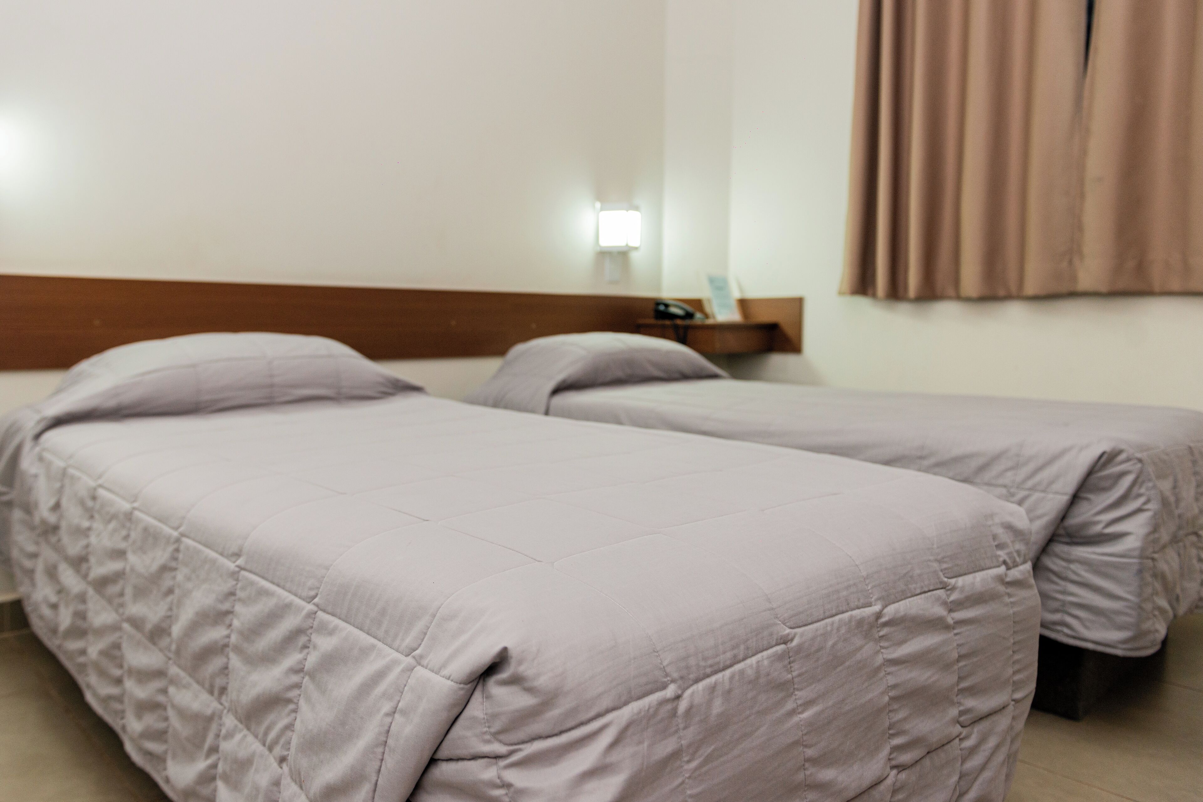 A hotel room with two beds, a nightstand, a lamp, and a window with curtains.