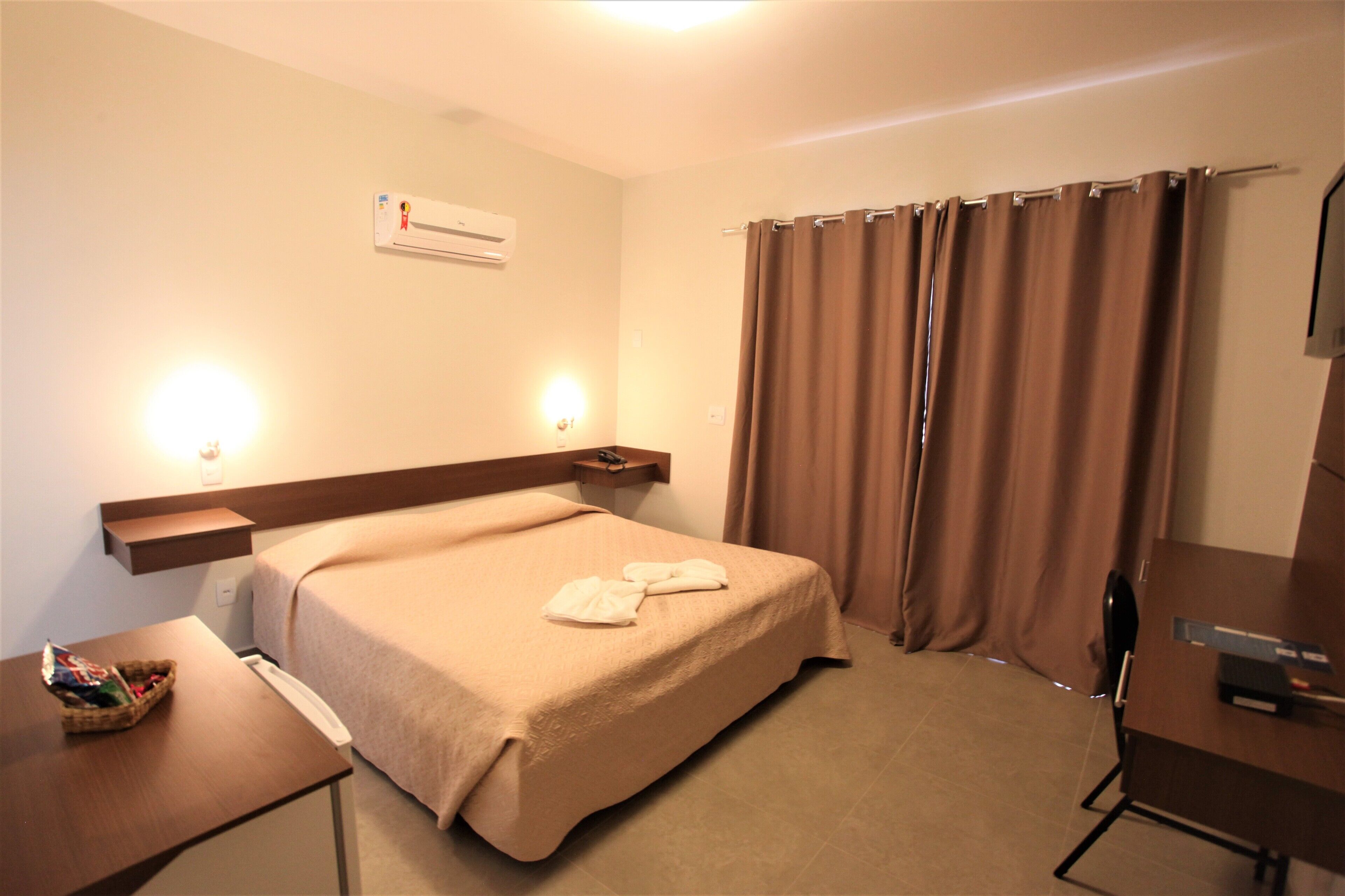 Quarto de hotel com cama, mesa de cabeceira, luminária e escrivaninha com cadeira.