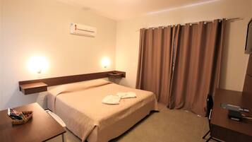 Quarto de hotel com cama, mesa de cabeceira, luminária e escrivaninha com cadeira.