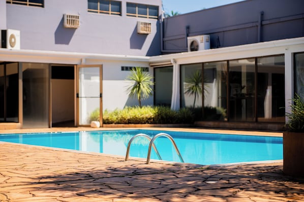 A swimming pool with a tiled deck, a glass door entrance, and a building with air conditioning units in the background. - Hotel Fernandão (Pouso Alegre)