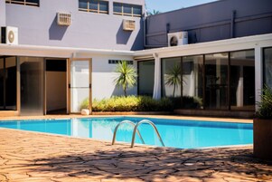 Una piscina con solárium azulejado, una entrada con puerta de cristal y un edificio con unidades de aire acondicionado al fondo.