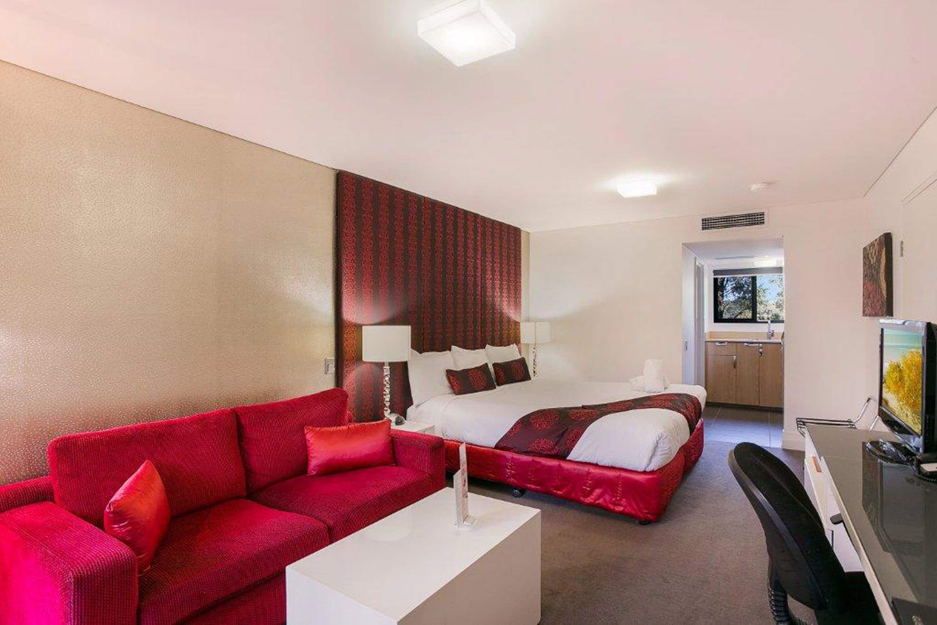 Habitación de hotel con un sofá rojo, una cama con ropa blanca, un escritorio con una silla y un televisor.