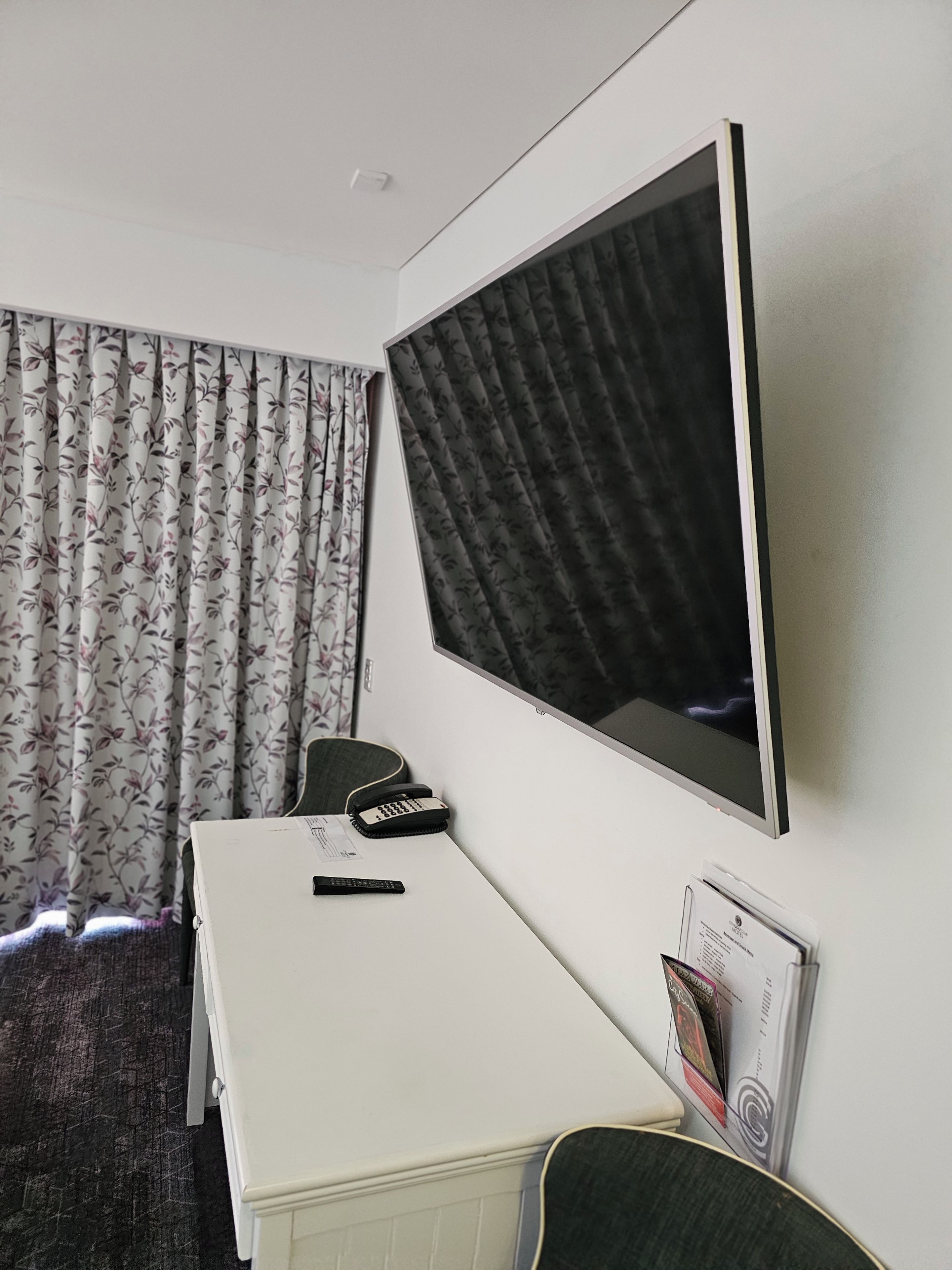 A hotel room with a bed, a desk, a chair, a flat-screen TV, a telephone, and a window with curtains.