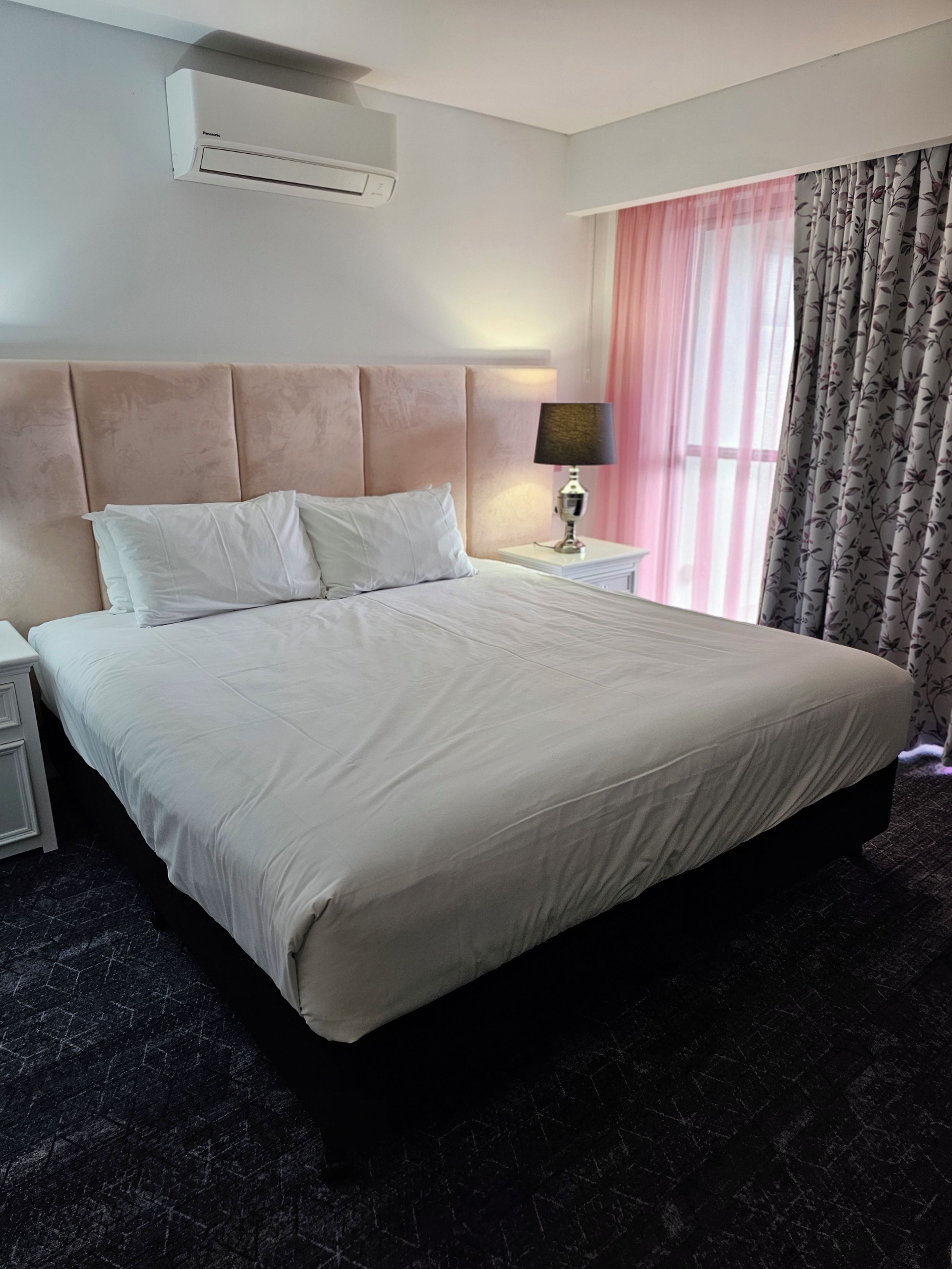 A hotel room with a large bed, a nightstand, a lamp, a window with curtains, and an air conditioning unit.
