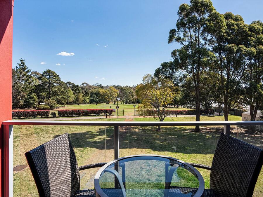 Un balcón con dos sillones de mimbre y una mesa de vidrio, con vista a un parque con árboles y un cielo despejado.
