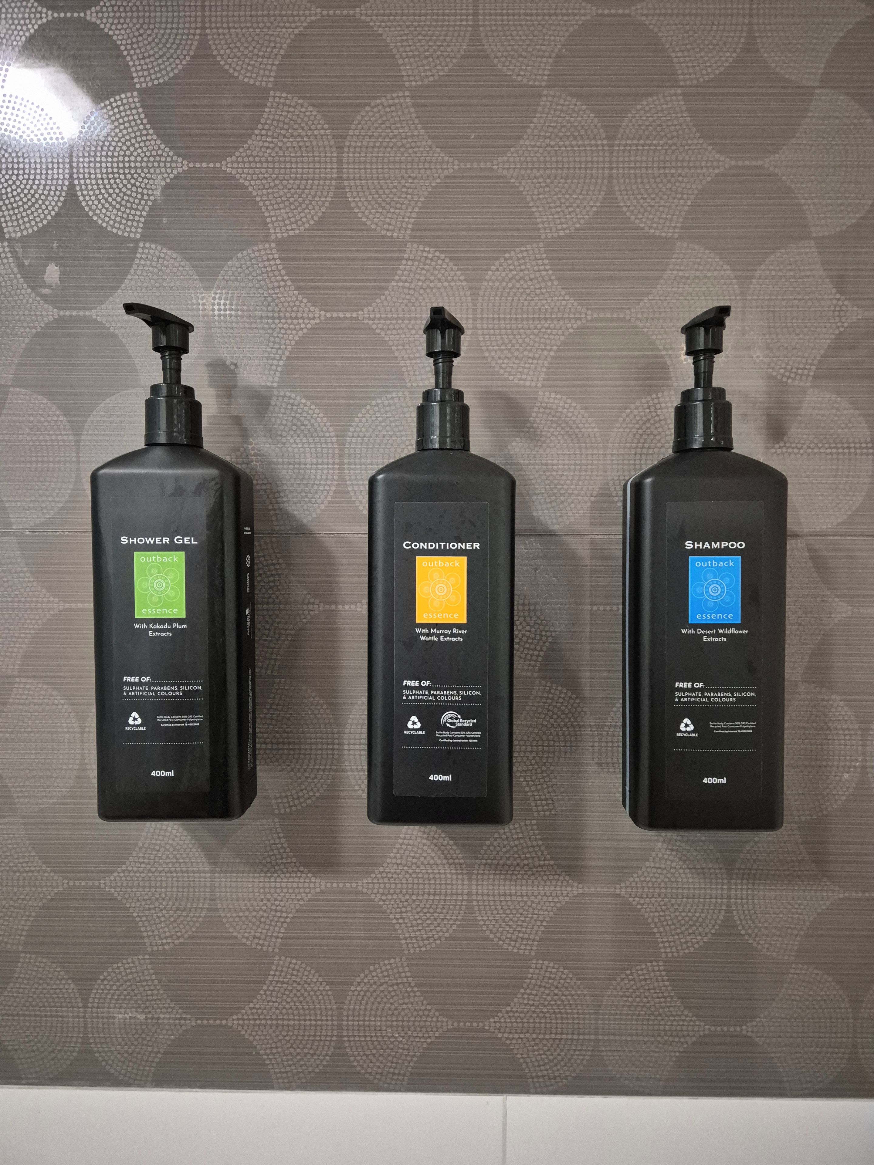 Drei schwarze Flaschen von Outback Essence Körperpflegeprodukten an einer Wand mit geometrischem Muster.