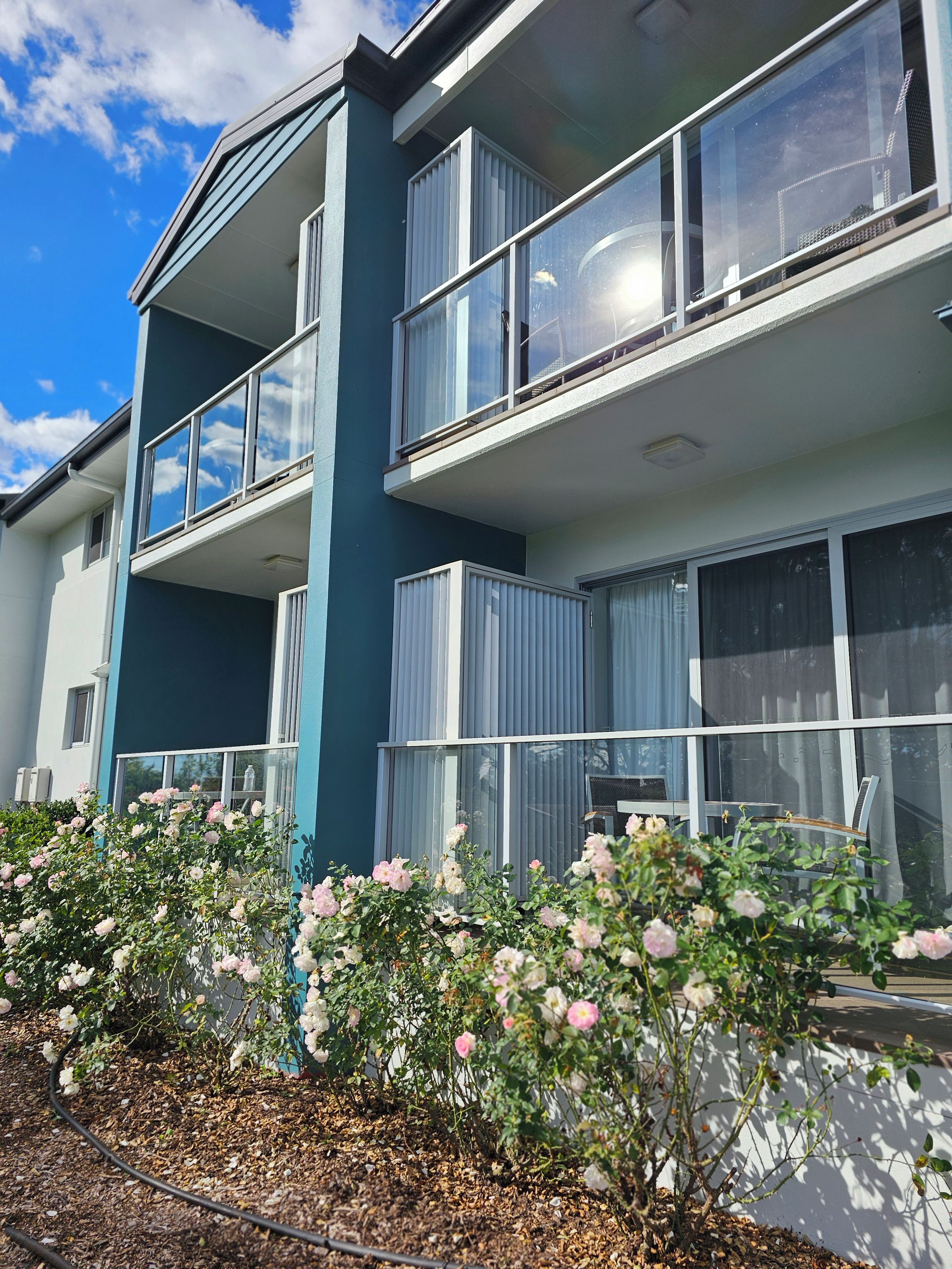 A modern two-story residential building with balconies, large windows, and a garden with flowering plants.