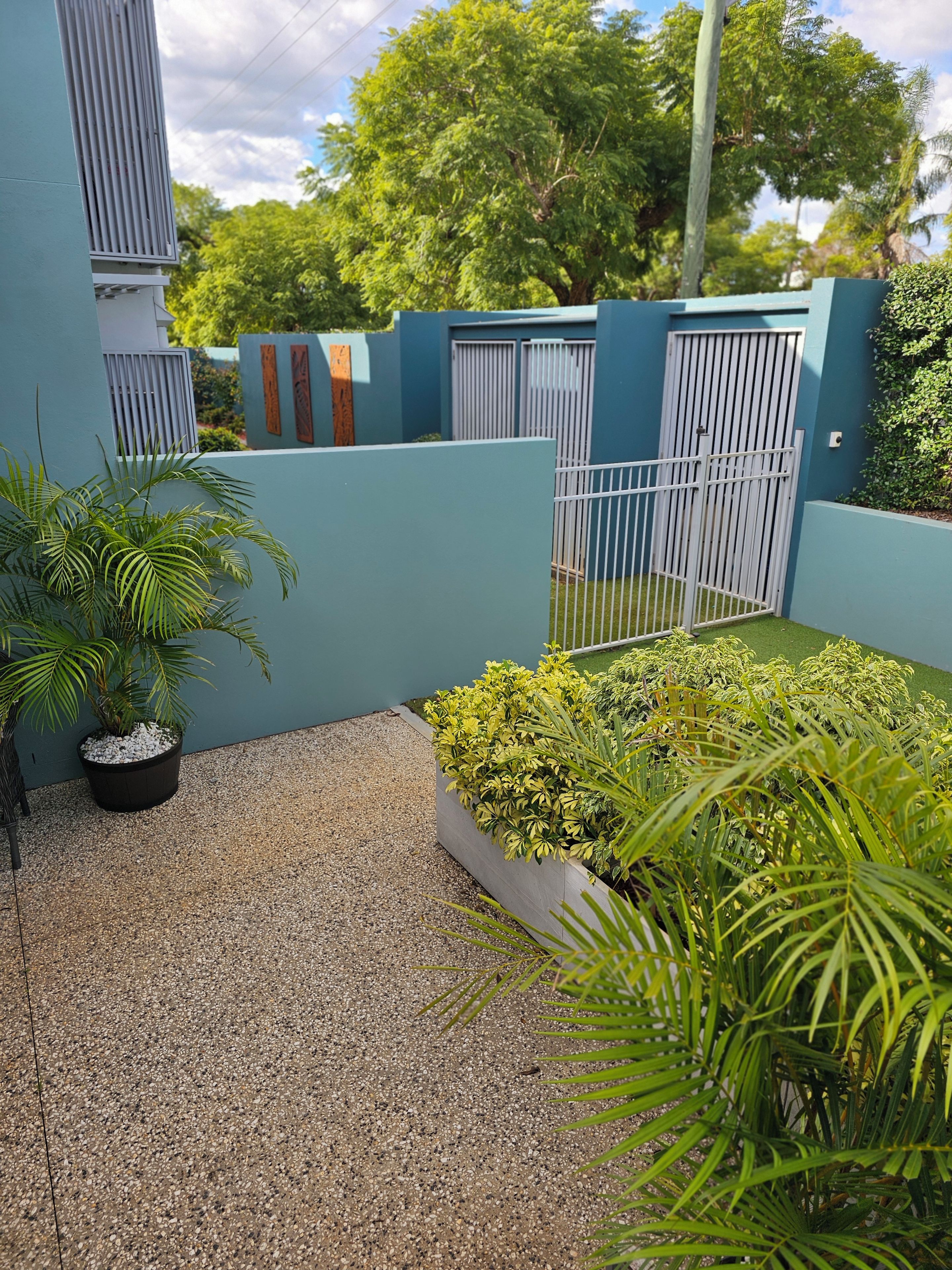Un patio con sendero de grava, plantas en macetas y una reja metálica.