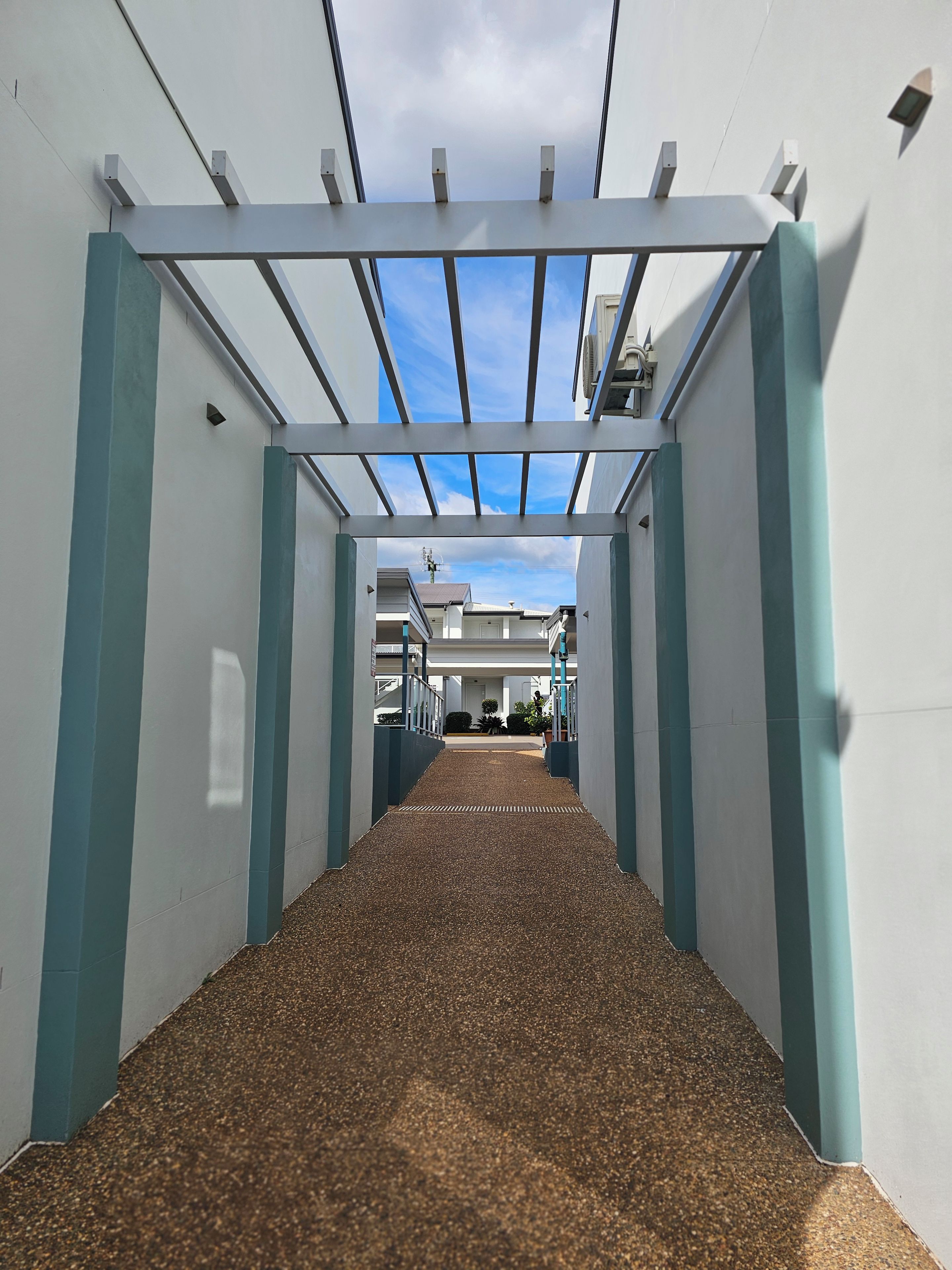 A corridor with a ceiling structure, a gravel floor, and a view of a building with a white facade and blue accents.