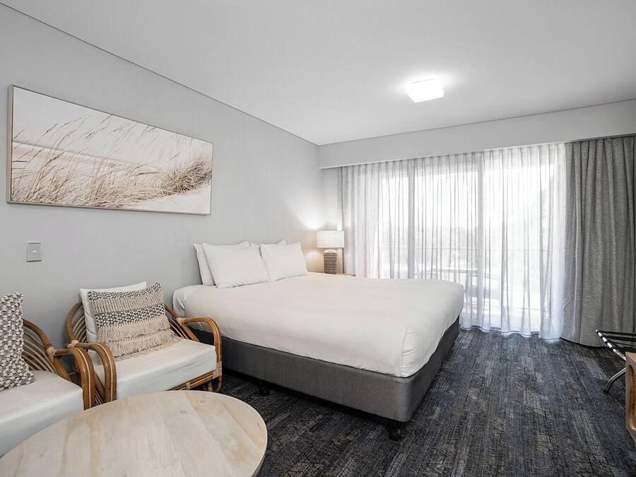 A hotel room with a large bed, a seating area with a chair and a small table, a large window with curtains, and a wall-mounted artwork.
