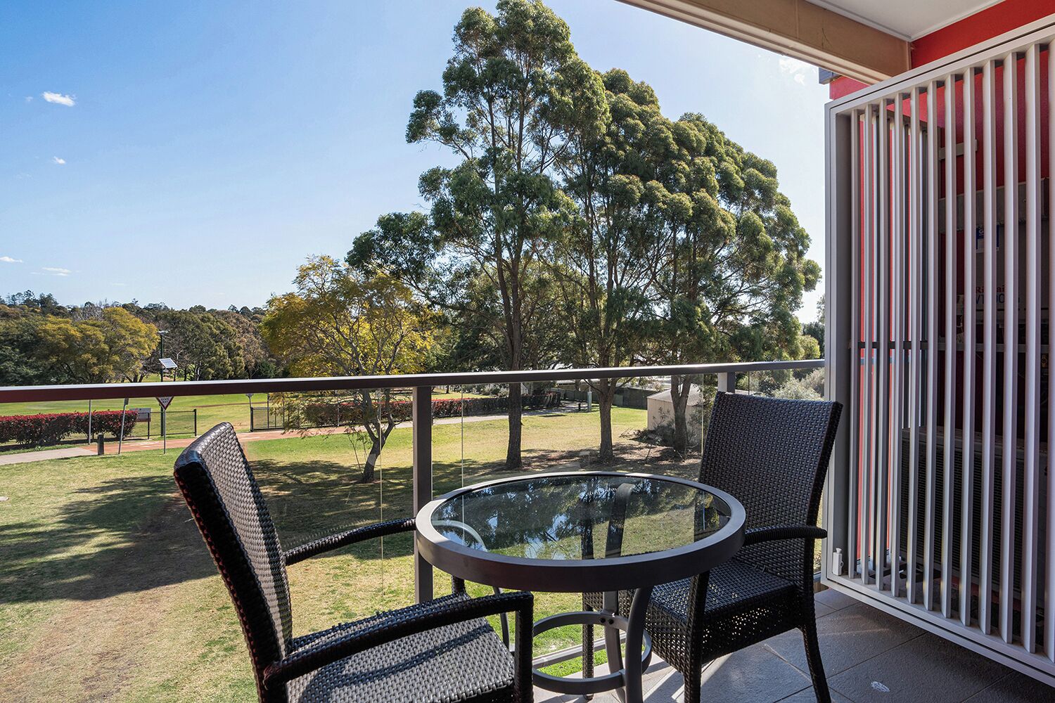 A balcony with a glass table and two wicker chairs, overlooking a grassy area with trees and a distant view of a golf course.
