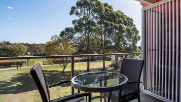 A balcony with a glass table and two wicker chairs, overlooking a grassy area with trees and a distant view of a golf course.
