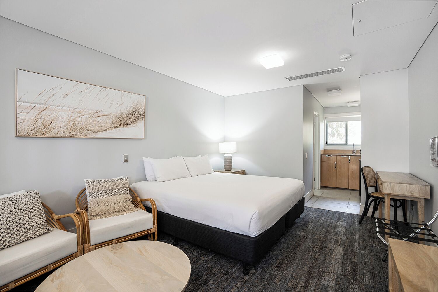 A modern hotel room with a large bed, a sitting area with a chair and a small table, a kitchenette, and a bathroom visible through an open door.