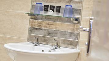 A bathroom with a white sink, two faucets, and a glass shelf above holding three bottles and a soap dispenser.