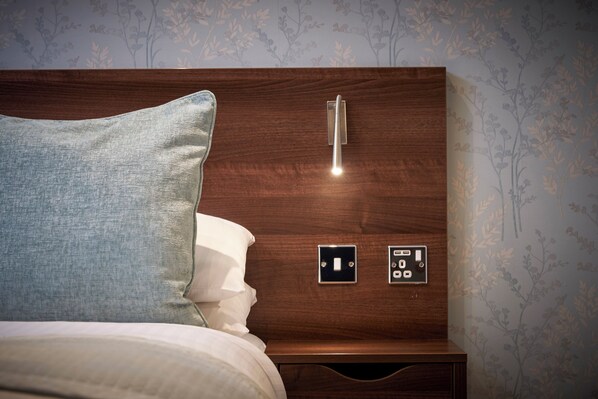 Un lit bien fait, avec un oreiller bleu et une tête de lit en bois dotée d’un interrupteur et d’un panneau de commande.