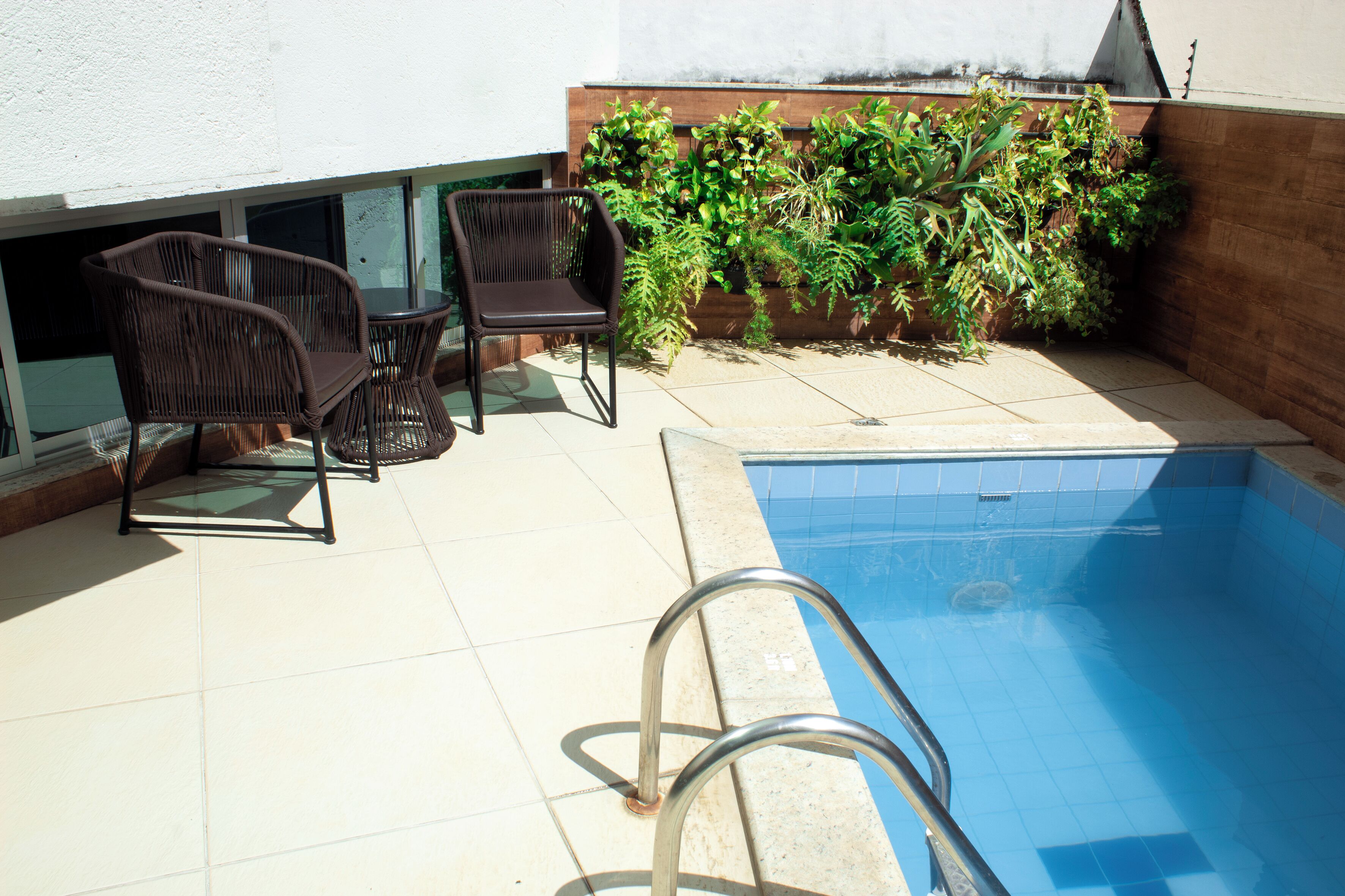 A swimming pool with a metal ladder, surrounded by a tiled patio area with wicker furniture and a planter with green plants.