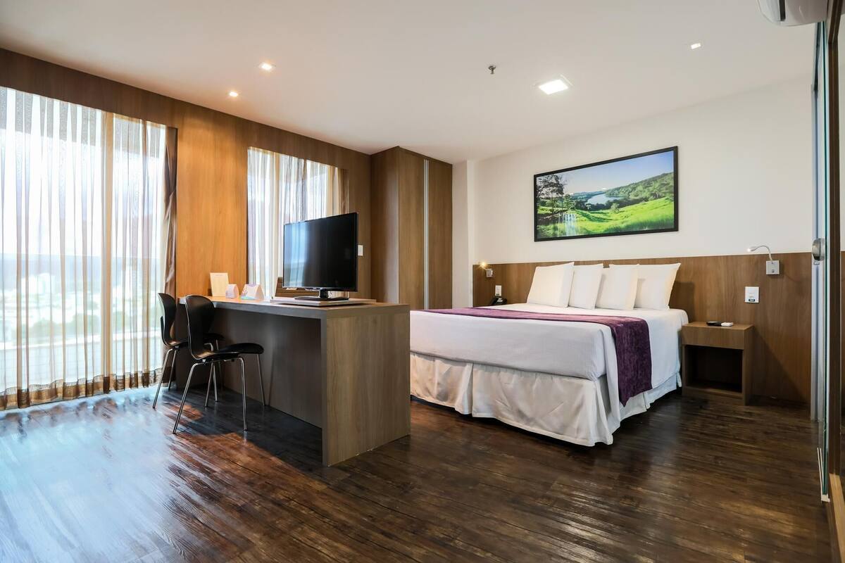 Una habitación de hotel con una cama, un escritorio con televisor, una silla y una ventana con cortinas.