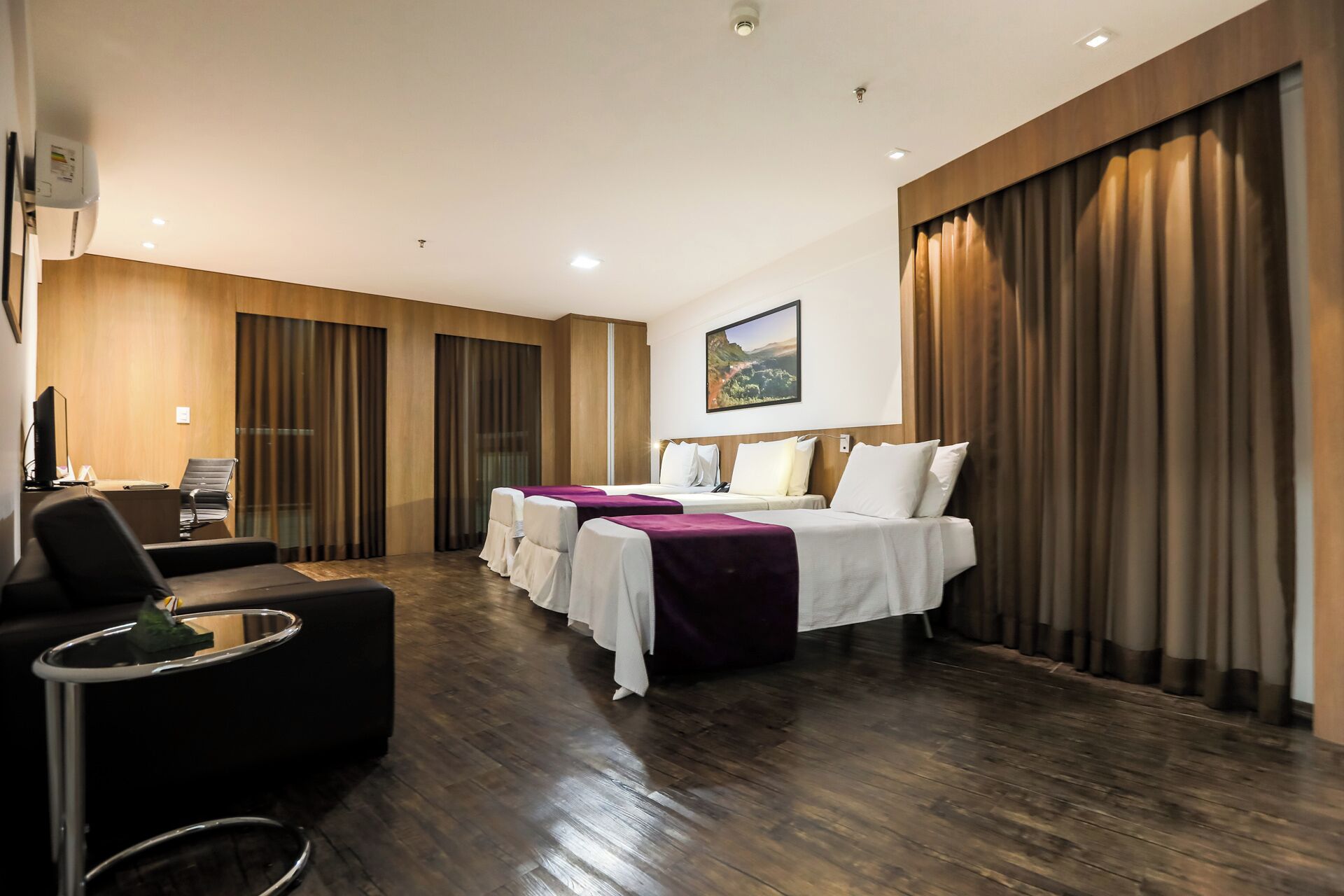A hotel room with two beds, a desk with a chair, a small table, and a large window with curtains.