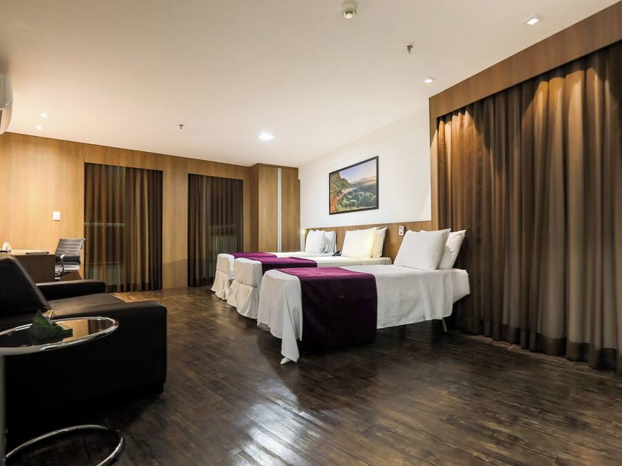 Quarto de hotel com duas camas, uma mesa com cadeira, uma mesinha e uma janela grande com cortinas.