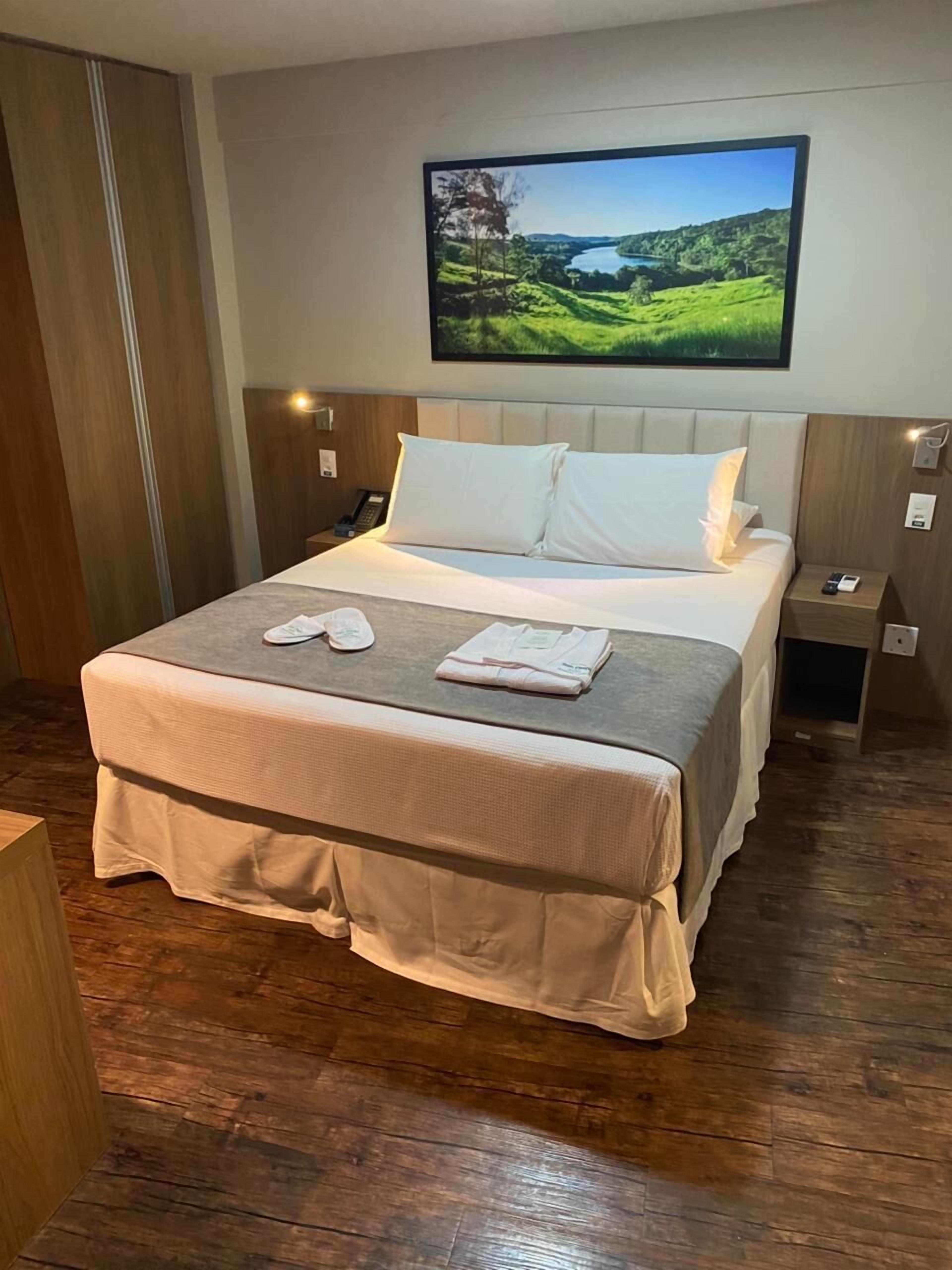 Un lit bien fait, avec une couette blanche et des oreillers, une table de chevet sur laquelle se trouvent un téléphone et une petite lampe, et un tableau représentant un paysage, accroché au mur.