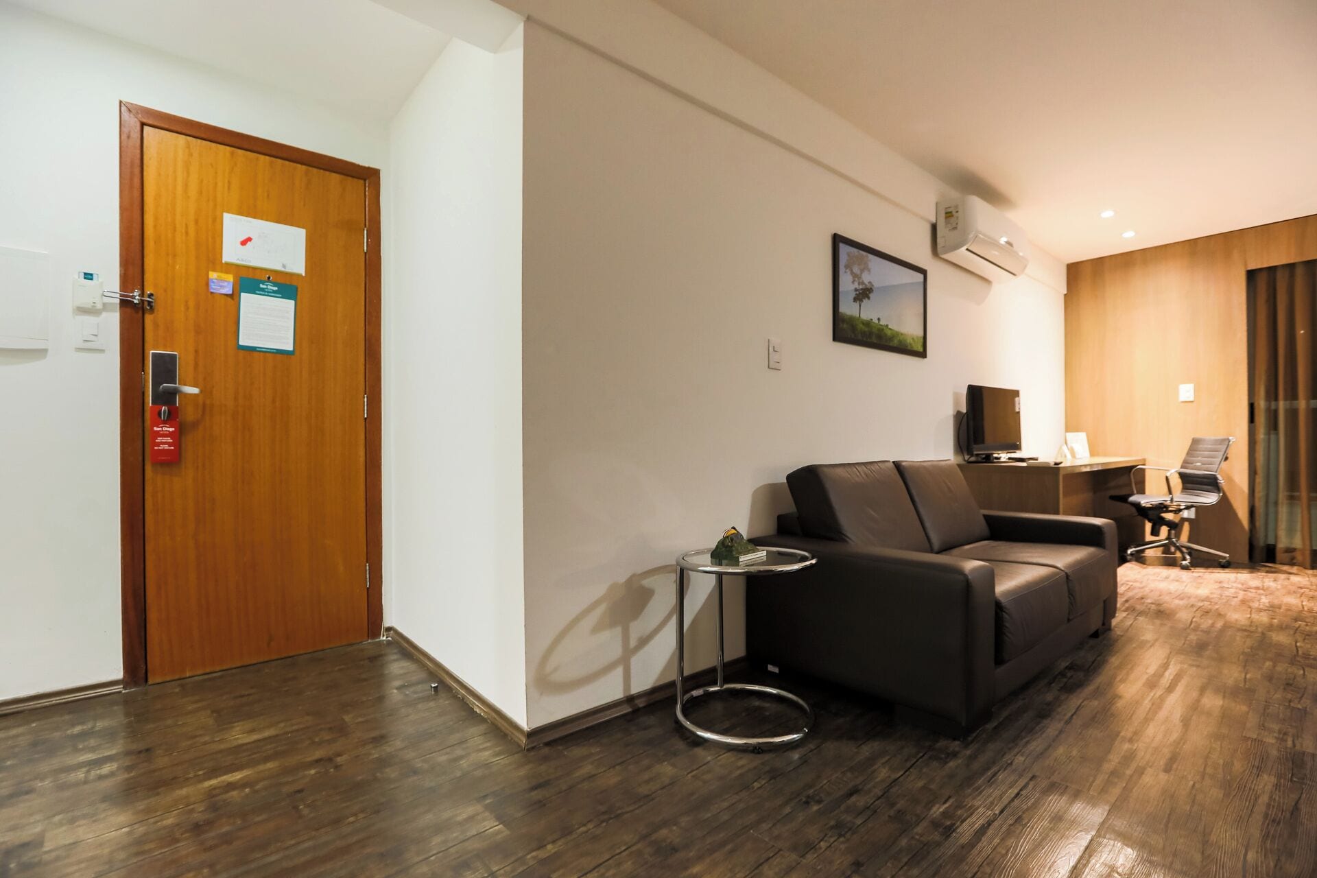 A modern hotel room with a wooden door, a black leather sofa, a small side table, and a television.