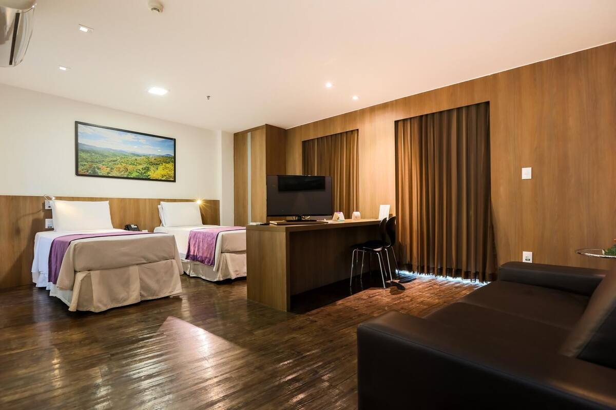 Habitación de hotel con dos camas, un escritorio con televisor, un sofá y un amplio ventanal con cortinas.