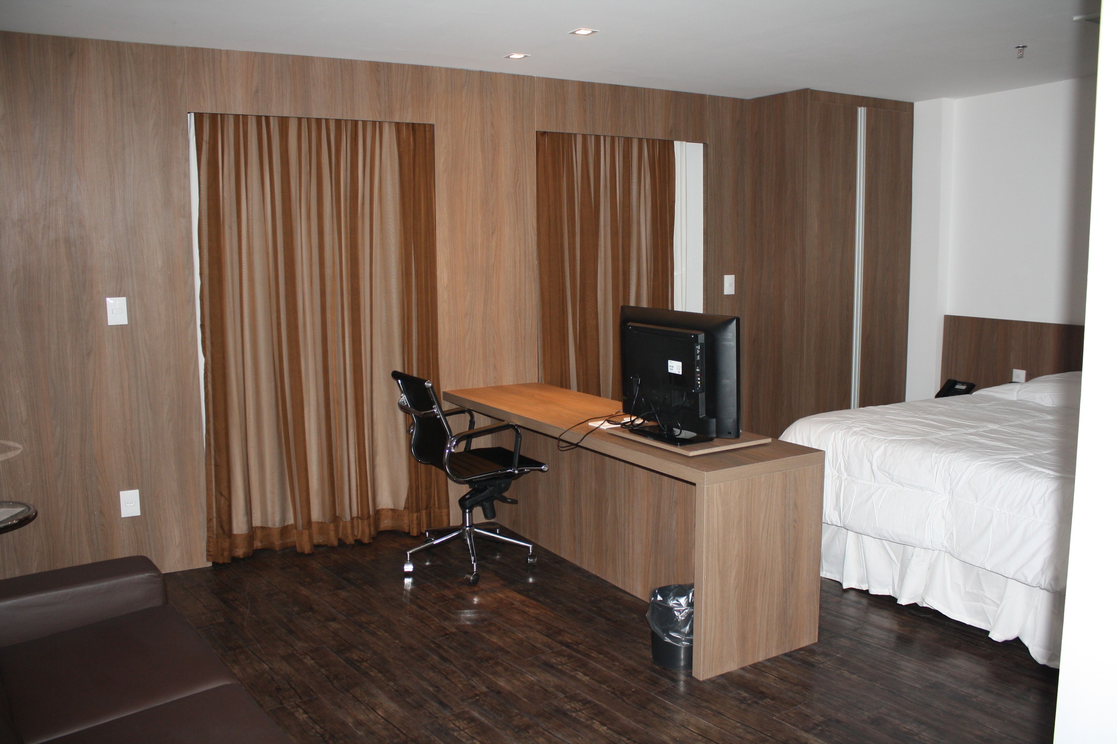 Quarto de hotel com escrivaninha de madeira, cadeira de escritório preta, cama com roupa de cama branca e uma janela grande com cortinas.