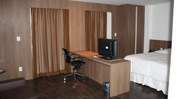 Quarto de hotel com escrivaninha de madeira, cadeira de escritório preta, cama com roupa de cama branca e uma janela grande com cortinas.