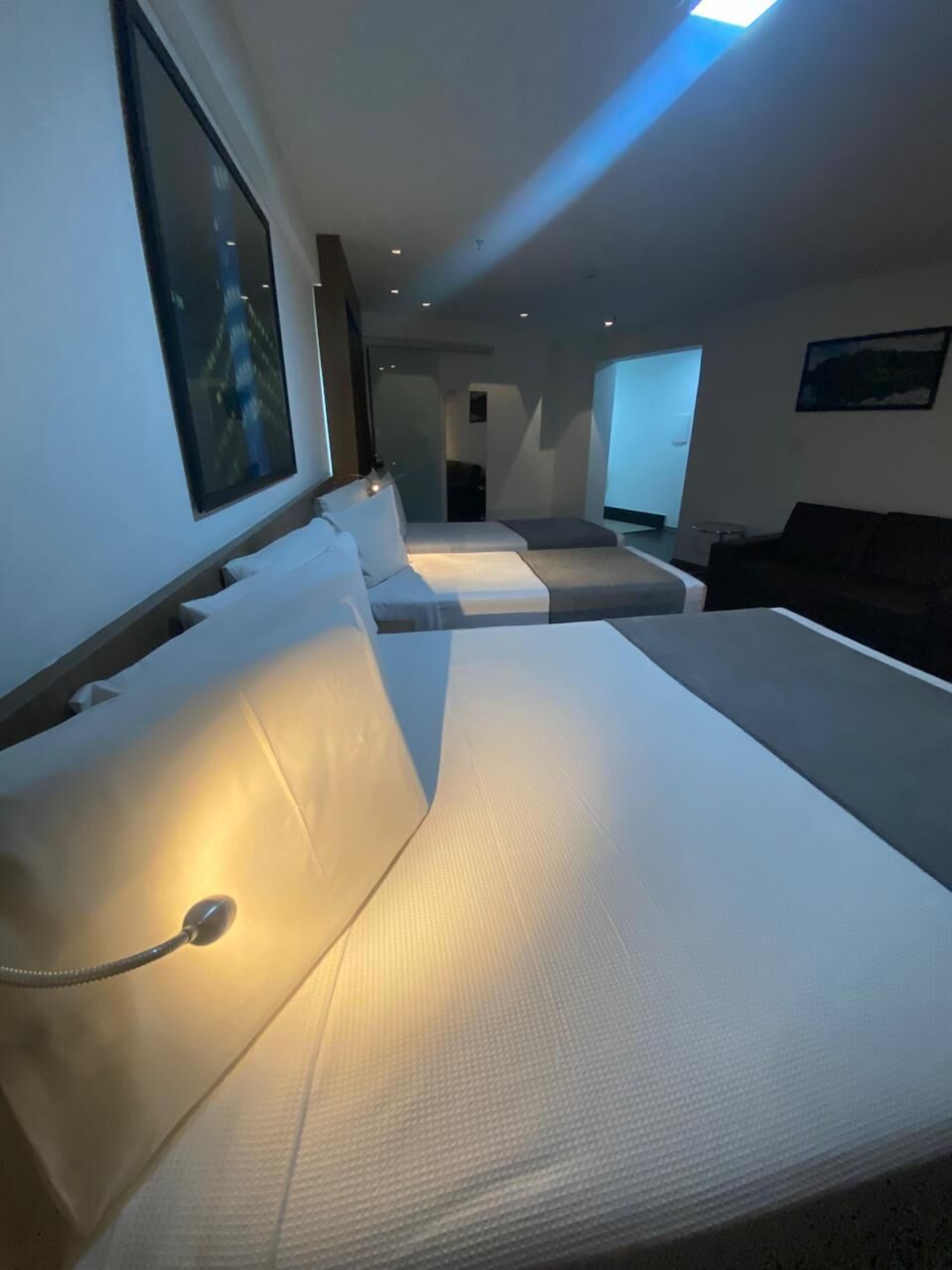 Une chambre d’hôtel moderne, de forme longue et étroite, comprenant une rangée de lits avec du linge de lit blanc et un lit simple doté d’une tête de lit sombre.