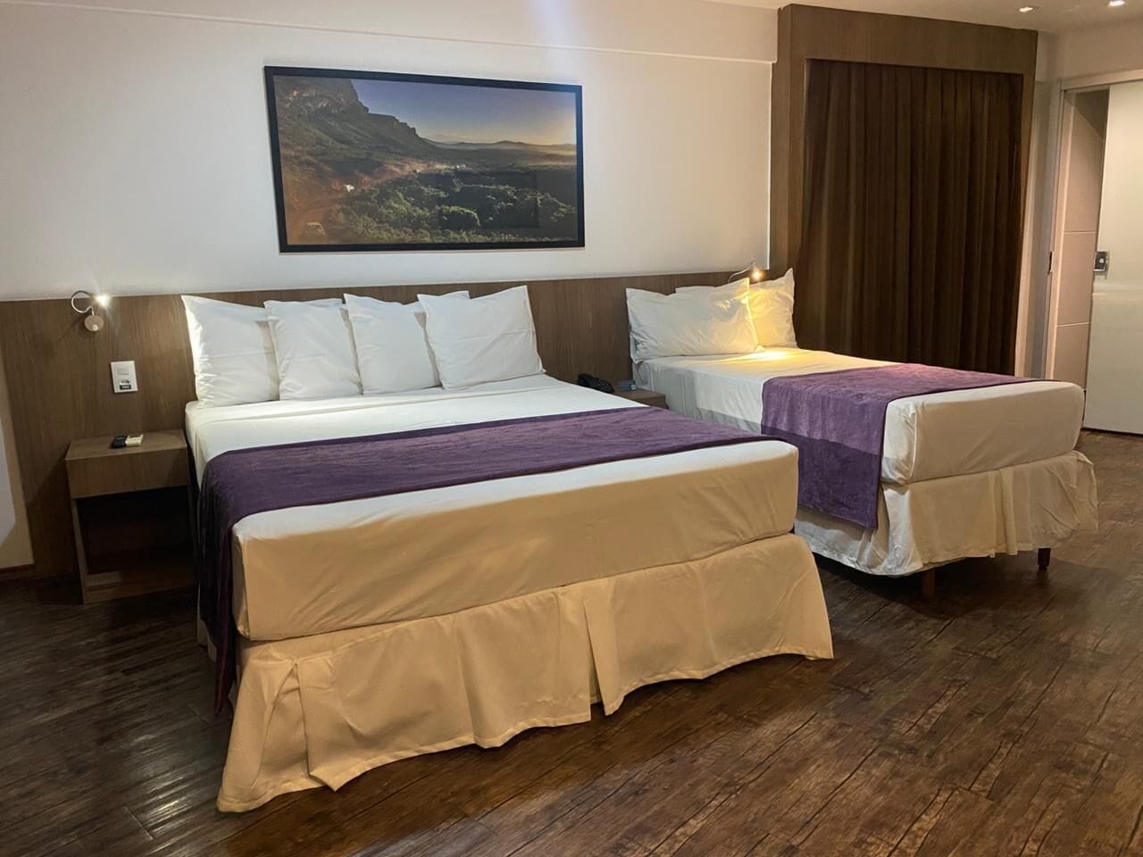 A hotel room with two beds, a wooden floor, a painting on the wall, and a curtain on the door.