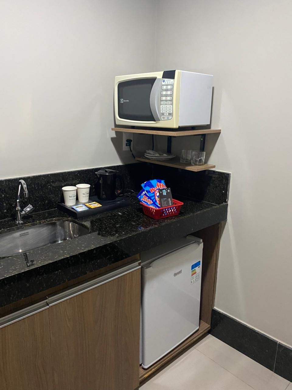 A compact kitchen with a microwave, a small refrigerator, a sink, and a countertop with a red basket containing snacks.