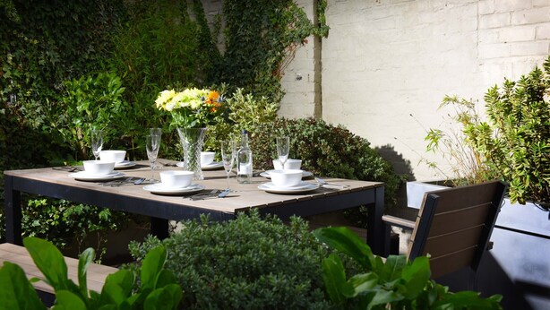 An outdoor dining area with a table set for four, surrounded by greenery and a white brick wall. - Arthouse (Canterbury)